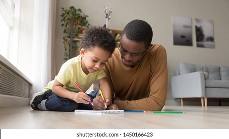 Loving Young African American Dad Lying On Floor With Little Son Drawing In Album At Home Together, Black Father Spend Time Learning With Preschooler Boy Child Paint Picture With Pencil On Paper