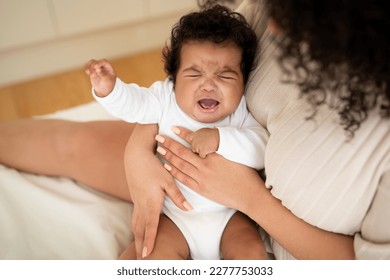 Loving young african american woman hold in her arms, calms crying cute baby, suffering from teeth growth on bed in light bedroom interior, top view. Love, mother and child at home, baby care - Powered by Shutterstock