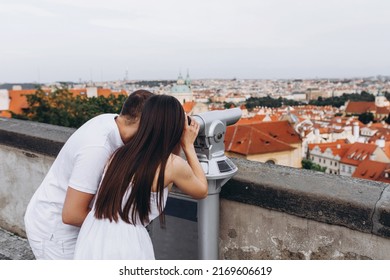 A loving young beautiful couple from Europe hugging in front of a beautiful view of the rooftops of the old houses in Prague, the couple dressed in white. The lovers look through binoculars at Prague.