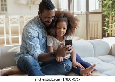 Loving young biracial dad sit on couch with cute little daughter watch funny video on smartphone together, happy caring african American father and small girl child use cellphone gadget at home - Powered by Shutterstock