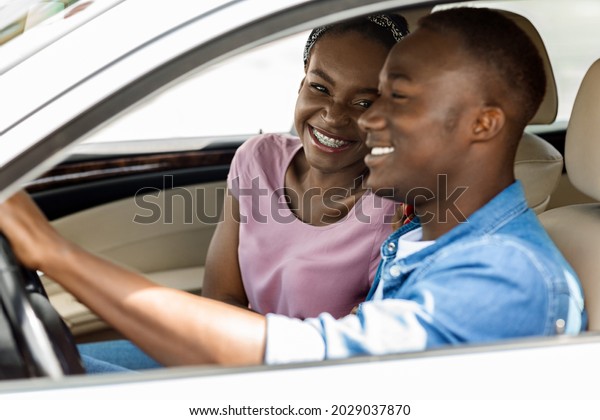 Loving young black man and woman sitting inside new\
auto, having conversation and laughing, having summer car trip to\
countryside, happy african american couple enjoying weekend\
together, side view