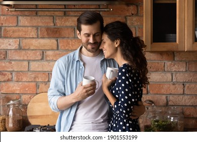 Loving Young Caucasian Couple Hug And Cuddle Drinking Coffee Or Tea In Morning In Cozy Home Kitchen. Happy Millennial Man And Woman Embrace Enjoy Family Weekend Time In Own House. Relation Concept.