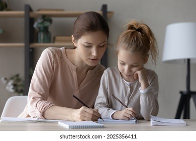 Loving young Caucasian mom help little daughter with homework assignment study write in notebook. Caring mother and small girl child handwrite take notes prepare home task. Education concept.