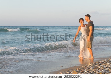 Loving young couple at the beach , in a late summer hazy day at dusk, wearing  a white dress and shorts, enjoying, going barefoot in the ocean water, getting wet, teasing and kissing one another.