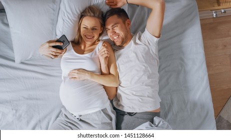 Loving Young Couple Spending Morning in Bed, Pregnant Young Woman Shows Her Partner Something on a Touchscreen Smartphone, Taking Selfie and Sharing Picture on Social Networks. - Powered by Shutterstock