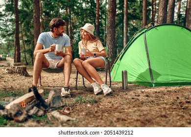A loving young couple went on a weekend for a picnic, sit near a campfire and a tent, drink coffee in a pine forest. Camping, recreation, hiking.
