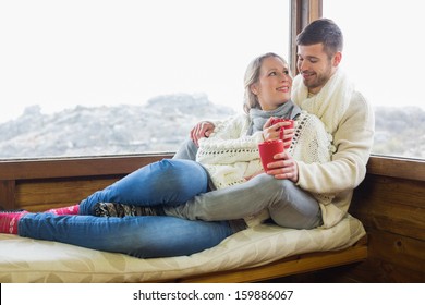 Loving Young Couple In Winter Clothing With Coffee Cups Sitting Against Cabin Window