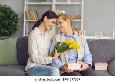 Loving Young Daughter Hugs And Gives Flowers And Gifts To Her Mature Mother In Honor Of Mother's Day. Older Woman And Her Daughter Look At Each Other With Tenderness And Love While Sitting On A Sofa.