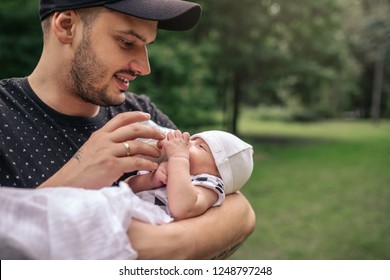 Loving Young Father Holding His Baby Boy In His Arms And Feeding Him With A Formula Bottle While Standing Outside In A Park