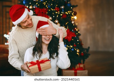 Loving young handsome man giving surprise gift to his wife, closing her eyes, celebrating Christmas at home - Powered by Shutterstock