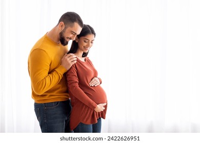 Loving young husband hugging his pregnant muslim wife from behind while they standing together near window at home, happy expectant caucasian family enjoying future parenting, copy space - Powered by Shutterstock