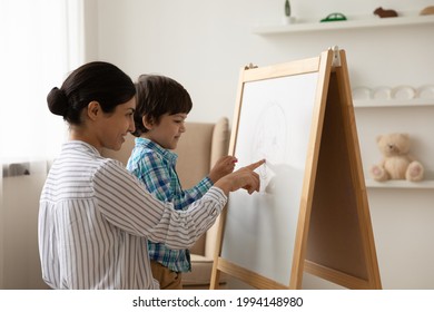 Loving young Indian mom and little ethnic son draw paint together on flip chart at home. Happy mixed race mother play learn study with small biracial boy child on whiteboard. Funny education concept.