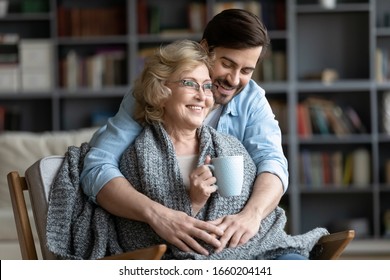 Loving young man take care of happy middle-aged mother sit in chair drinking tea enjoying family weekend in living room, thankful grown-up adult son hug show gratitude and affection to mature mom