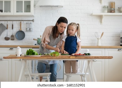 Loving young mom and cute small preschooler daughter have fun preparing food in kitchen together, caring mom teach little girl child cooking, help chop vegetables for salad, family weekend concept