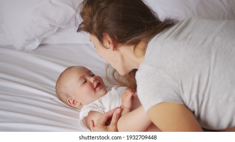 Loving Young Mommy Hugging Soothing Adorable Sweet Baby Girl Lying In Bed. Smiling Caring Mother And Cute Little Infant Child Girl Cuddling In Bedroom. Mum And Child Tender Moments.