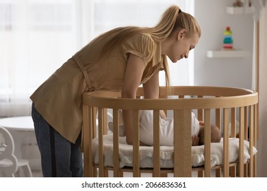 Loving young mother putting sleeping small baby in wooden crib bed, tender moment, caring mom holding cute newborn child infant standing in cozy children bedroom nursery, motherhood concept