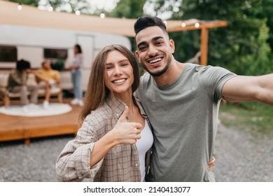 Loving young multiracial couple taking selfie near motorhome, resting with their friends outdoors. Arab guy and his Caucasian girlfriend enjoying summer camping trip, making mobile photo - Powered by Shutterstock