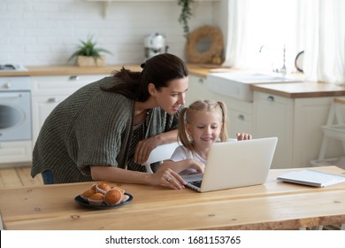 Loving young mum and little preschooler daughter sit at kitchen table using laptop together, caring mother help small girl child teaching learning online on computer, kids and technology concept