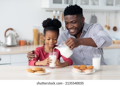 Loving young single black father pouring into glass fresh milk for his cute little daughter, sitting together at cozy kitchen, having snack or healthy breakfast together, drinking milk, eating cookies - Powered by Shutterstock