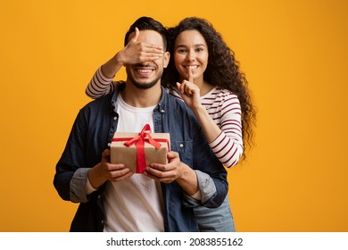 Loving Young Woman Covering Boyfriend's Eyes And Surprising Him With Gift, Cheerful Middle Eastern Lady Showing Shh Sign, Greeting Her Husband With Birthday Or Valentines Day Over Yellow Background