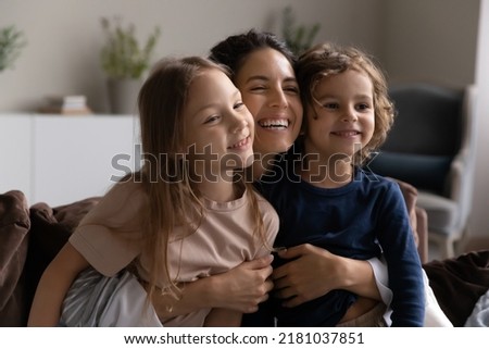 Loving young woman cuddling her preschool son and daughter, family smiling looking aside sit on sofa in living room, close up view. Concept of happy motherhood, unconditional mothers love, adoption