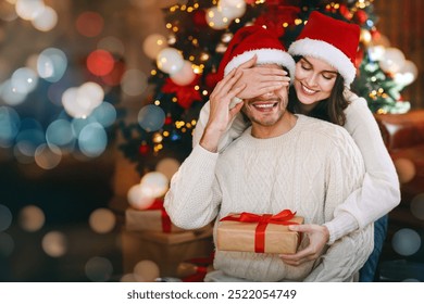 Loving young woman giving surprise gift to her husband, closing his eyes, celebrating Christmas at home - Powered by Shutterstock