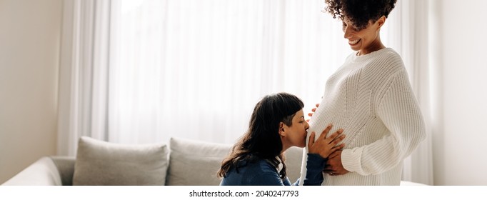 Loving young woman kissing her wife's pregnant belly at home. Young lesbian woman showing love and affection to her unborn baby. Happy young lesbian couple expecting a baby.