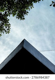 Low angle abstract building corner. Low angle view of a sharp, modern dark building corner pointing toward a cloudy blue sky with green tree leaves framing the top.
