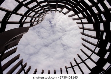 Low angle abstract view looking straight up through the circular opening of a wooden slat roof structure towards a cloudy blue sky.