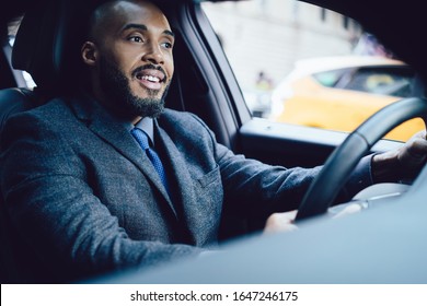 Low Angle Of African American Pleased Entrepreneur In Formal Clothes Looking Away And Smiling While Driving Luxurious Black Automobile In City