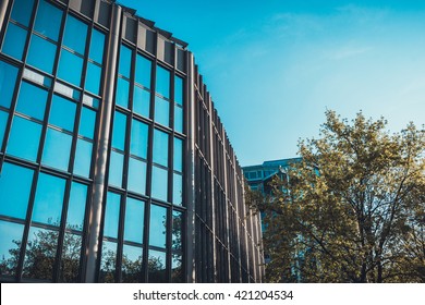 Low Angle Architectural View Of Low Rise Modern Office Building In Urban Setting With Green Deciduous Trees On Sunny Day With Blue Sky In Munich, Germany