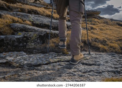 Low angle back view of hiker in leather boots walking on rocky mountain trail with trekking poles during autumn trip with scenic landscape and dramatic sky on background.