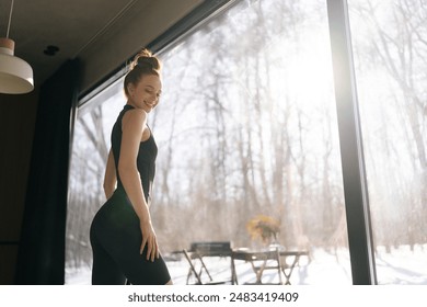 Low angle back view of smiling fitness female posing at home standing by window with beautiful snowy winter nature view on sunny day. Attractive successful woman coach in sportswear with closed eyes.
