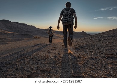 Low angle back view of two hikers with backpacks walking on rocky desert terrain during sunset adventure for summer travel exploration and active lifestyle in remote wilderness.