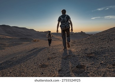 Low angle back view of two hikers with backpacks walking on rocky desert terrain during sunset adventure trip for summer travel exploration and active lifestyle in remote wilderness.
