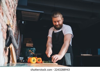 Low angle of bearded male chef in t shirt with apron cutting cucumbers and pepper on chopping board and looking at camera while cooking in kitchen at home