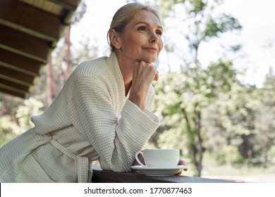 Low angle of calm mature lady enjoying view of nature from balcony while drinking coffee - Powered by Shutterstock