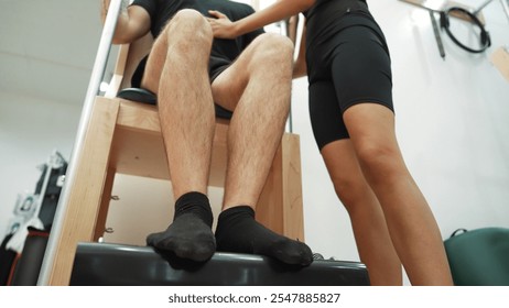 Low angle camera of pilates instructor assisting a client in a leg press exercise on a Wunda Chair for improving core engagement, and balance in a gym. Healthy man posing footwork on chair. Habituate. - Powered by Shutterstock