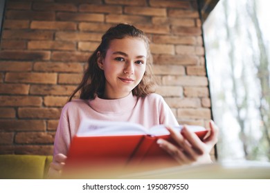 Low Angle Of Charismatic Lady In Casual Wear Looking At Camera While Reading Interesting Book In Red Cover In Modern Cafe