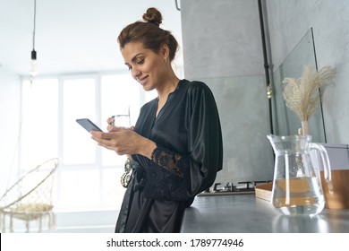 Low Angle Of Cheerful Young Female In Black Dressing Gown Using Cell Phone And Quenching Thirst In Kitchen