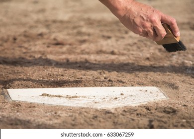 Low Angle Close Up Of Baseball Umpire Brushing Dirt Off Home Plate With Small Black Bristle Brush In Afternoon Sunlight