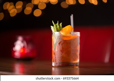 Low Angle Close Up Of Gourmet Old Fashioned Craft Cocktail Of Sweet Bourbon Whiskey Garnished By Orange Peel And Rosemary Sprig On Table Of Red Leather Diner Booth With Blurry Restaurant Background