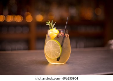 Low angle close up of ice cold modern gourmet craft cocktail of gin and tonic soda garnished by lemon slice and rosemary sprig sprinkled by juniper berries on bar with blurry restaurant bar background