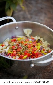 Low Angle Close Up Large Metal Cooking Pot Of Bowtie Pasta Salad Seasoned With Italian Spices And Raw Ingredient Of Banana And Bell Pepper And Red Onion Mixed With Green Olives On Weather Wood Deck 