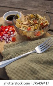 Low Angle Close Up Lunch Scene Of Bowtie Pasta Salad In Glass Bowl Seasoned With Italian Spices And Raw Ingredient Of Bell Pepper And Red Onion On Wood Cutting Board With Green Towel Napkin With Fork