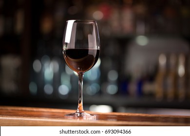 Low Angle Close Up Perspective Of Crystal Clear Wine Glass With Traditional Round Goblet Shape Filled With Dark Red Wine And Slim Stem On Wood Counter Top Bar With Blurry Restaurant Background Scene