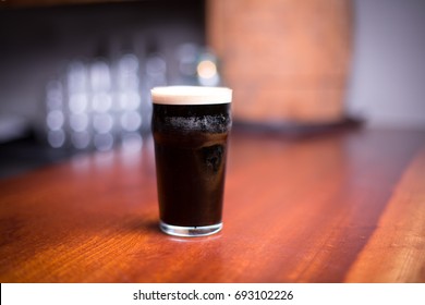 Low Angle Close Up Perspective Of Traditional Tumbler Pint Shape Beer Glass Filled With Dark Malt Stout Nitrogen Porter With Foam Head On Wood Counter Top Bar With Blurry Restaurant Background