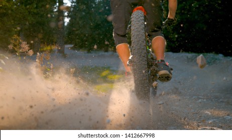 LOW ANGLE, CLOSE UP, DOF: Bright sunbeams shine on a part of a gravel path as the downhill cyclist rides past the camera and through the forest. Man brakes while riding e-bike down an empty trail.