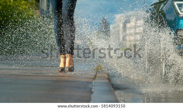 LOW ANGLE, CLOSE UP, DOF: Unrecognizable young\
woman wearing high heels gets splashed with water as inconsiderate\
driver drives their car into a deep puddle at the side of the empty\
asphalt road.