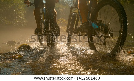 LOW ANGLE CLOSE UP: Two unrecognizable persons riding new mountain bikes across the sunlit stream and splashing water. Bright sun rays illuminate the river as men ride bikes through the dark forest.